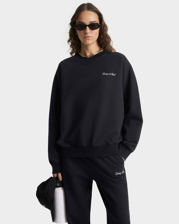 Person wearing a black sweatshirt and pants with a logo, holding a white cup on a light gray background