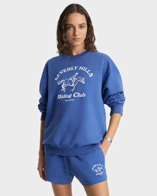 Woman wearing a blue 'Beverly Hills Riding Club' sweatshirt and shorts on a white background