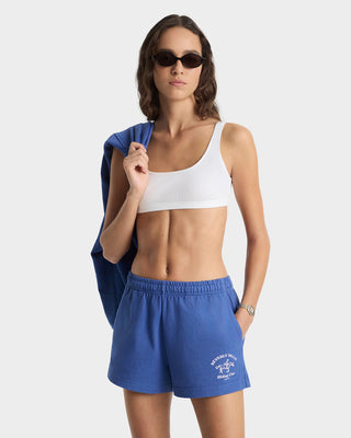 Woman wearing a white sports bra and blue shorts with a logo, holding a blue jacket.