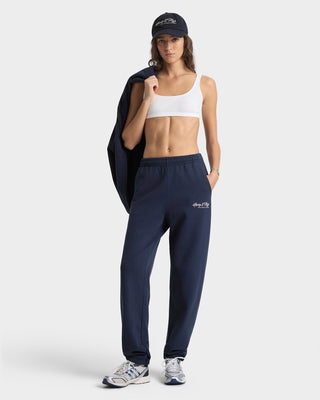 Woman wearing navy jogger pants and a white sports bra, holding a jacket on a white background