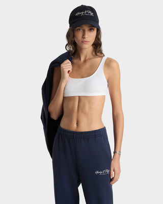 Woman wearing a white sports bra and navy pants with a matching cap on a light gray background