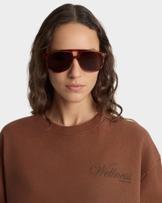 Person wearing brown sunglasses and a brown sweatshirt with 'Wellness' text on a white background