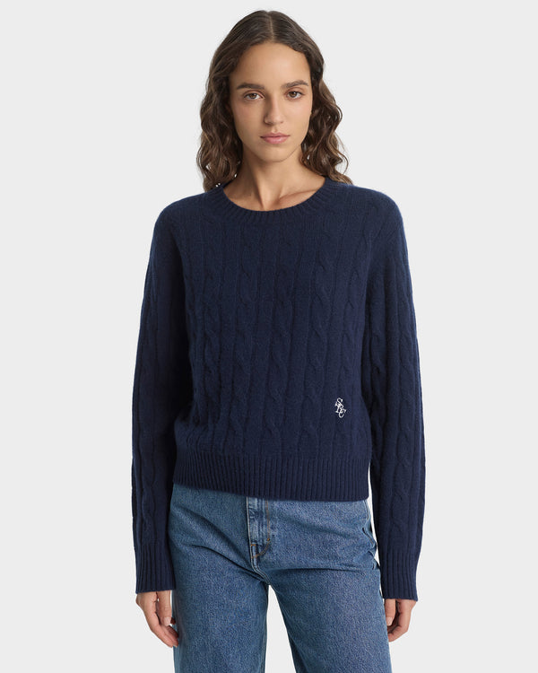 Woman wearing a navy blue cable knit sweater with a brand logo on a white background