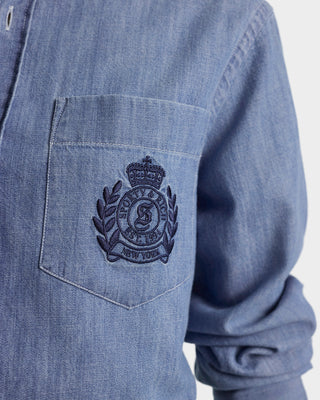 Blue denim shirt with a embroidered logo on the chest pocket against a white background