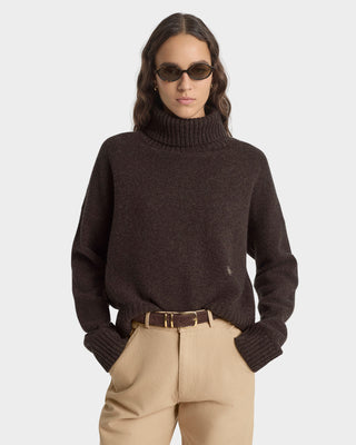 Person wearing a dark brown turtleneck sweater and beige pants on a light gray background