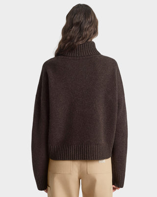 Person wearing a brown turtleneck sweater and beige pants on a light gray background