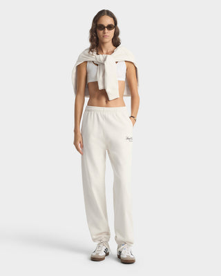 Woman wearing a white sleeveless top and pants with a logo, standing against a plain background.