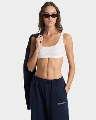 Woman wearing a white sports bra and navy sweatpants with a brand logo, holding a navy jacket.
