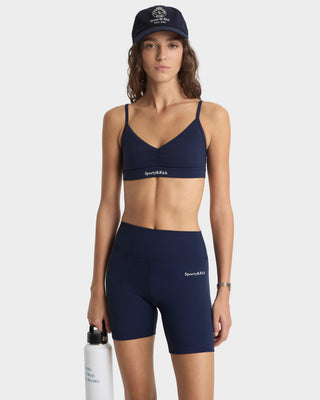 Woman wearing navy sports bra and shorts with visible branding, holding a white water bottle on a white background.