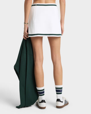 Person wearing a white tennis skirt with green trim, holding a green jacket, and wearing white sneakers with black stripes.