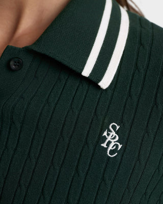 Dark green cable knit sweater with white trim collar and logo on a neutral background