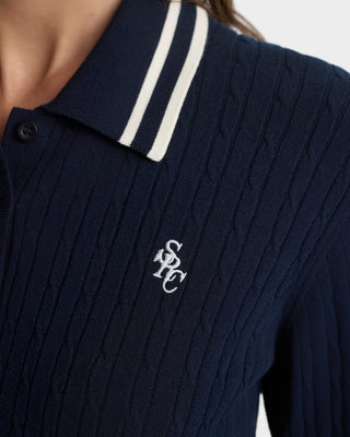 Navy blue cable knit sweater with white collar and logo on a light gray background