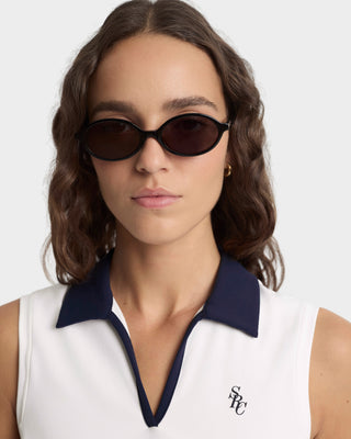 Woman wearing dark sunglasses with a white sleeveless top and navy collar.