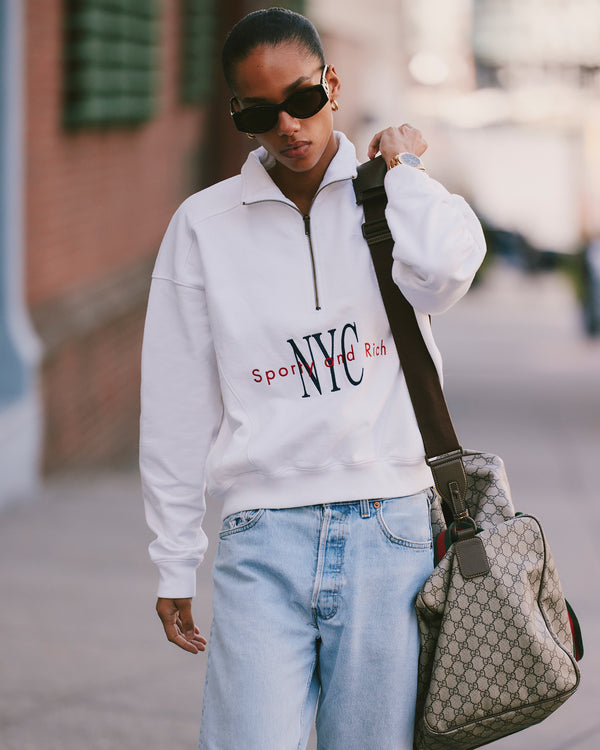 Person in sunglasses and a NY Minute Quarter Zip - White/Navy sweatshirt, made in Portugal from 100% cotton, walking with a large Gucci bag and wearing light blue jeans.
