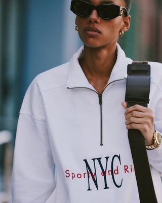 Person wearing sunglasses and a white NY Minute Quarter Zip - White/Navy sweatshirt with a black strap over their shoulder.