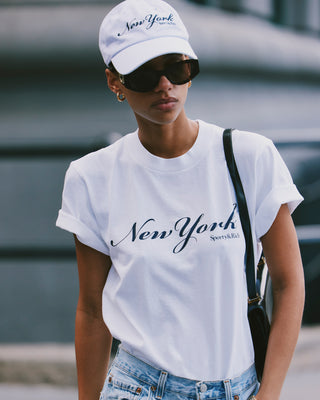 Woman in sunglasses wears a white New York T-Shirt - White/Navy and cap outdoors, carrying a black bag. Made from 100% cotton, this tee is shrink free for lasting comfort and fit.