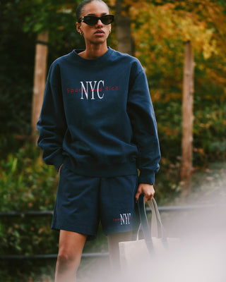 Woman in navy NYC sweatshirt and NY Minute Gym Short - Navy/White, 100% cotton heavy jersey with drawstring waistband, wearing sunglasses and holding a tote bag outdoors with greenery behind.