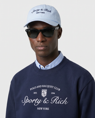 Man wearing a navy sweatshirt with 'Sporty & Rich' branding, light blue cap, and sunglasses on a plain background