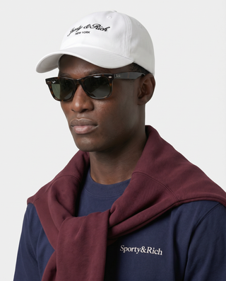 Man wearing a white cap, sunglasses, and a burgundy hoodie with 'Sporty & Rich' branding.