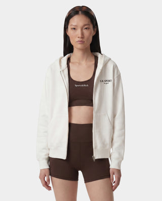 Woman wearing a white hoodie and brown sports bra and shorts on a white background