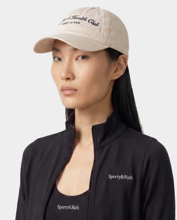 Person wearing a beige cap and black jacket with 'Sporty&Rich' branding on a white background