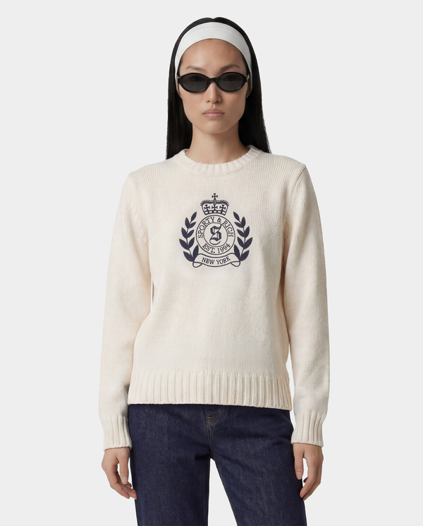 Woman in sunglasses and headband, wearing the NY Crest Knitted Sweater - Ecru/Navy (100% cotton, embroidered logo), blue jeans, and flip-flops, standing against a white background.