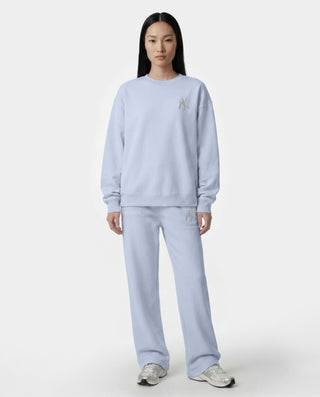 A model stands against a light gray studio background wearing a matching light blue sweatshirt and sweatpants set with small silver embroidered details on the chest and thigh.