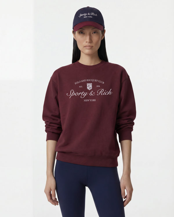 Person wearing a burgundy sweatshirt with 'Sporty & Rich' branding and a matching cap on a white background