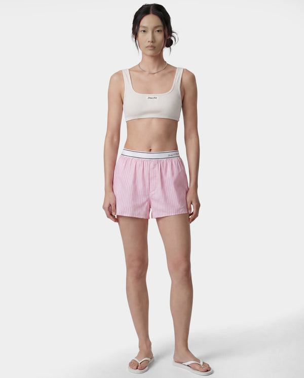 A model stands against a light gray studio background wearing a matching two-piece loungewear set consisting of a cream-colored crop top with a small black logo and pink-and-white vertically striped shorts with a white elastic waistband.