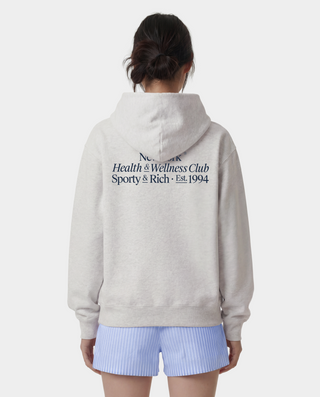 A model shown from the back wearing a light gray hoodie with black text on the back reading 'New York Health & Wellness Club Sporty & Rich Est. 1994', paired with light blue and white striped shorts against a light gray background.