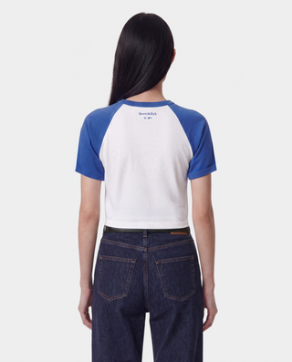 A woman wearing a blue and white Los Angeles Dodgers baseball helmet, sunglasses, a blue and white raglan crop top with 'Dodgers' written on the front, and white athletic shorts, standing on a baseball field.