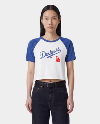 A woman wearing a blue and white Los Angeles Dodgers baseball helmet, sunglasses, a blue and white raglan crop top with 'Dodgers' written on the front, and white athletic shorts, standing on a baseball field.