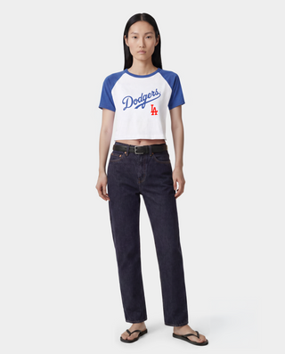 A woman wearing a blue and white Los Angeles Dodgers baseball helmet, sunglasses, a blue and white raglan crop top with 'Dodgers' written on the front, and white athletic shorts, standing on a baseball field.