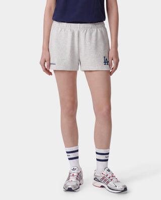 A woman outdoors on a baseball field wearing a light gray Los Angeles Dodgers sweatshirt and matching shorts, holding a tan baseball glove, with a navy cap featuring an LA logo.