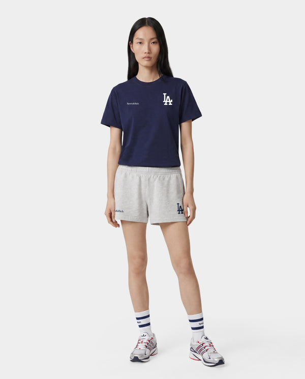 A woman outdoors on a baseball field wearing a light gray Los Angeles Dodgers sweatshirt and matching shorts, holding a tan baseball glove, with a navy cap featuring an LA logo.