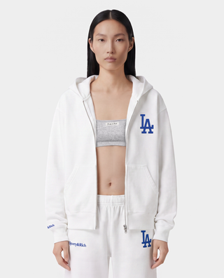 The LA Serif Zip Hoodie - White/Navy features a white zip-front design, crafted from 100% cotton, with a blue LA logo on the left chest and a front pocket, displayed against a plain background.