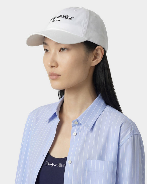 Woman wearing a white cap with branding and a light blue striped shirt on a plain background
