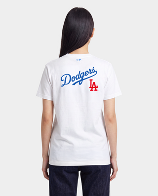 A woman on a baseball field wearing a white Los Angeles Dodgers t-shirt from the back, featuring the team's script logo and 'LA' logo in blue and red, paired with blue athletic shorts and a white cap.