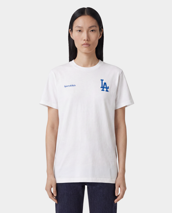 A woman on a baseball field wearing a white Los Angeles Dodgers t-shirt from the back, featuring the team's script logo and 'LA' logo in blue and red, paired with blue athletic shorts and a white cap.