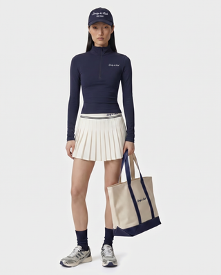 Woman wearing a navy long-sleeve top, white pleated skirt, and holding a tote bag with a visible brand logo.