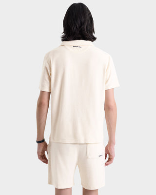 Model shown from the back wearing Eden Script Terry Short in Cream/Navy, a pair of 100% cotton terry shorts, styled with a matching cream short-sleeve shirt against a plain backdrop.