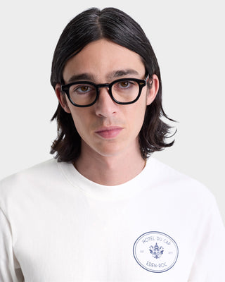 A person with long dark hair and black glasses wears the Kennedy Crest T-Shirt in White/Navy, made in Portugal from 100% cotton, featuring a Hotel Du Cap logo, facing forward.