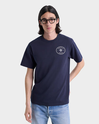 Person with long hair and glasses wears an oversized Kennedy Crest T-Shirt - Navy/White (100% cotton) and light jeans, standing against a plain background.