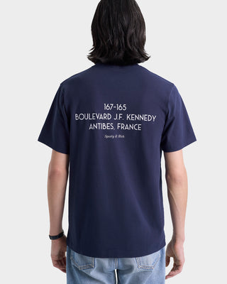 Person wearing the Kennedy Crest T-Shirt in navy/white, made in Portugal, featuring white BOULEVARD J.F. KENNEDY, ANTIBES, FRANCE text on the back.