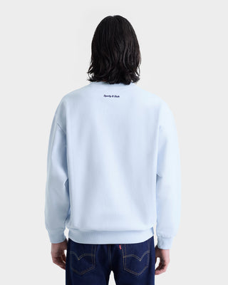 Person with long dark hair wears the Eden Crest Crewneck - Ciel/Navy, an oversized light blue sweatshirt made in Portugal from 100% cotton, paired with dark jeans and shown from the back.