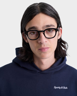 Man with shoulder-length dark hair, black glasses, and a navy oversized Eden Crest Hoodie - Navy/White by Sporty & Rich. Made in Portugal from 100% cotton.