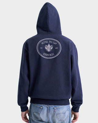 Wearing the Eden Crest Hoodie - Navy/White, made in Portugal from 100% cotton with the Hotel du Cap Eden-Roc logo on the back, paired with light blue jeans.