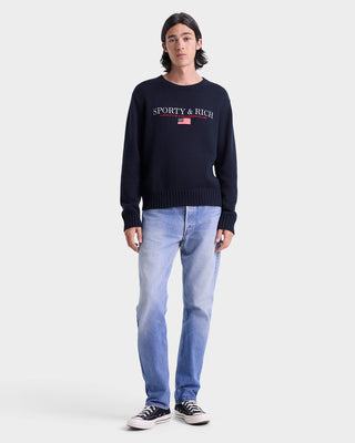 Person wearing a navy blue 'Sporty & Rich' sweater and light blue jeans on a white background