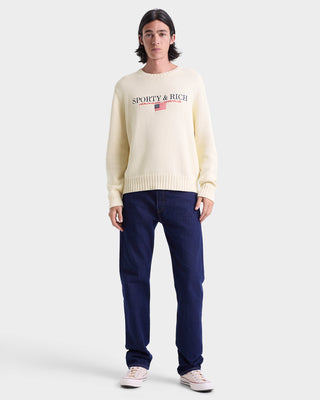 Person wearing a cream sweater with 'Sporty & Rich' logo and dark blue jeans on a white background