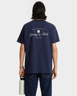 A person faces away from the camera, holding a bottle and wearing the Syracuse T-Shirt in navy with white text on the back.